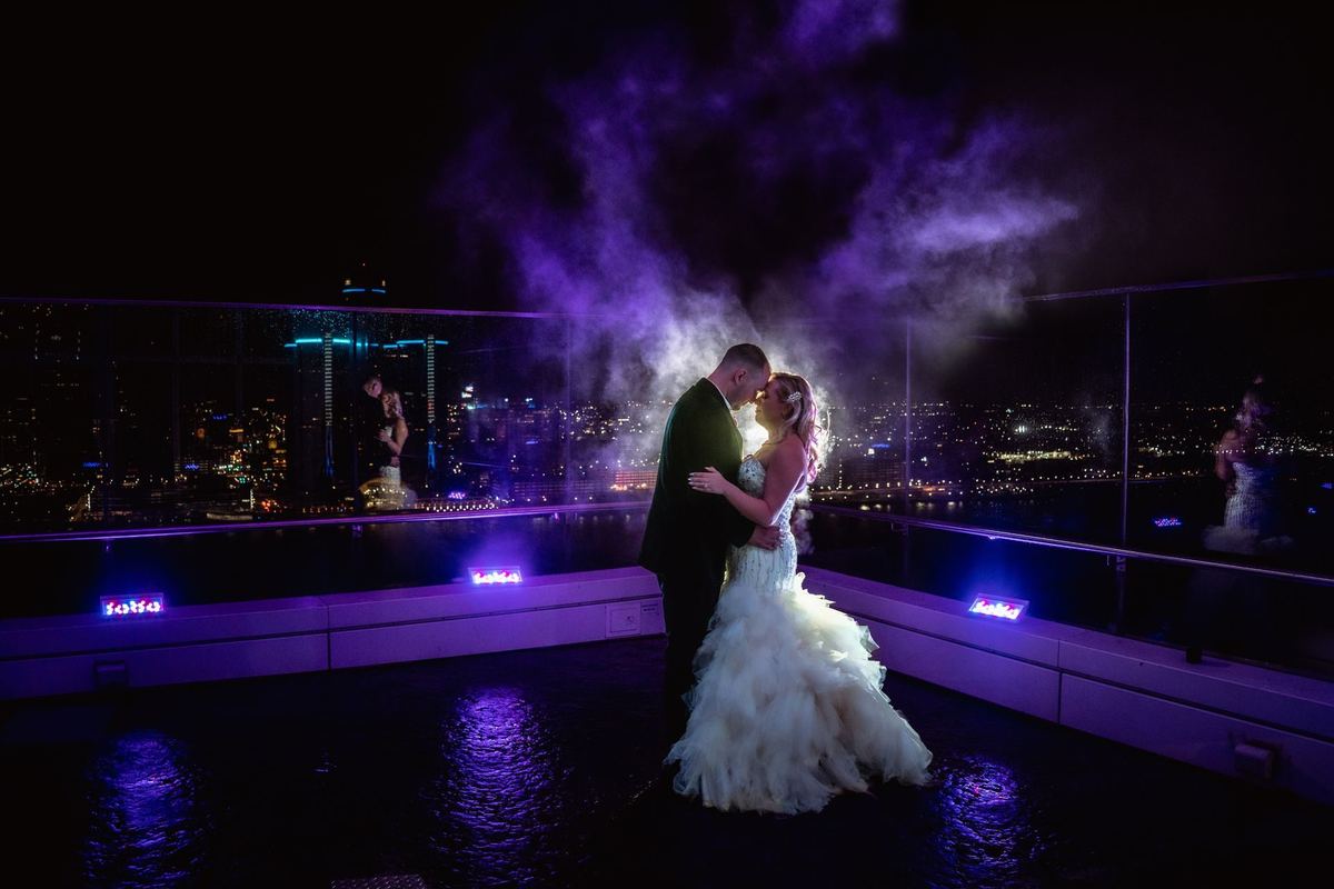 Bride and groom embracing on a rooftop terrace, backlit by purple smoke and city lights at the end of the night