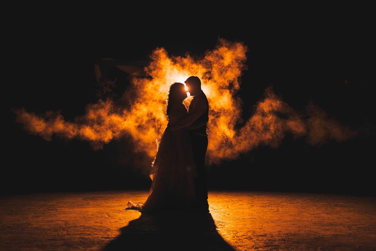 Bride and groom silhouetted against glowing orange smoke in an end-of-night backlit wedding portrait