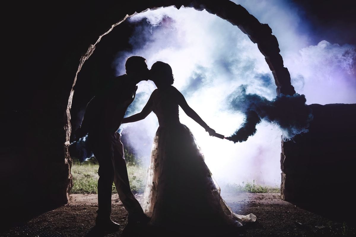 Couple kissing under a stone archway, backlit by white smoke in an end-of-night wedding portrait