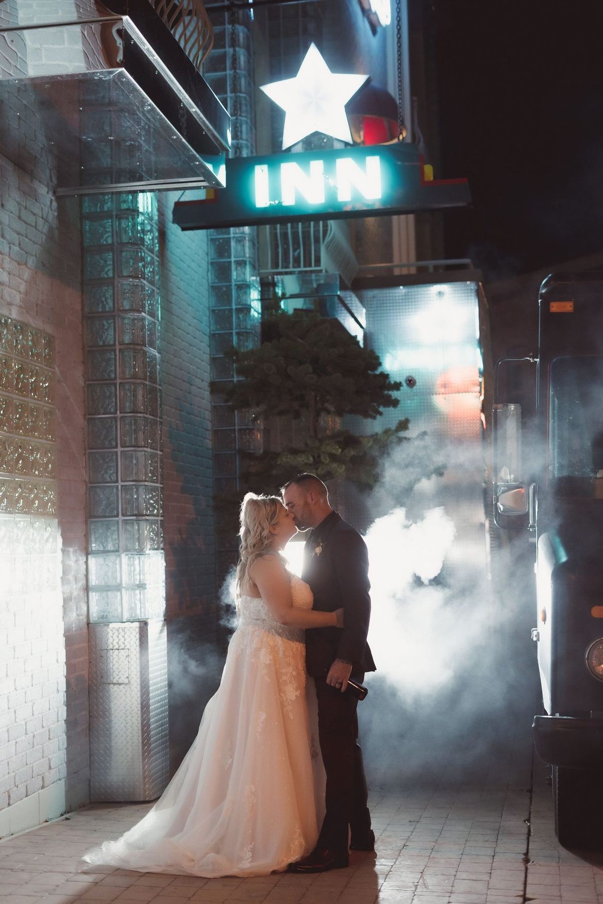 Bride and groom backlit by smoky light beside a neon star sign in an end-of-night portrait near Retro Suites