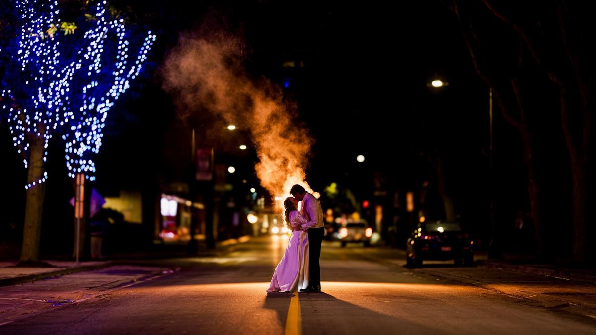 Bride and groom on a city street at night, backlit by smoke and blue tree lights, wedding portrait