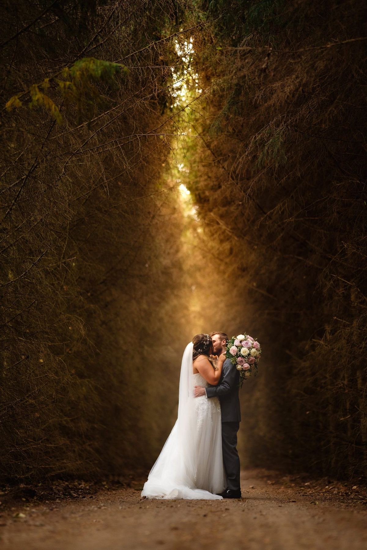 Bride and groom embracing in a dark tunnel of trees, backlit by a glowing light behind them
