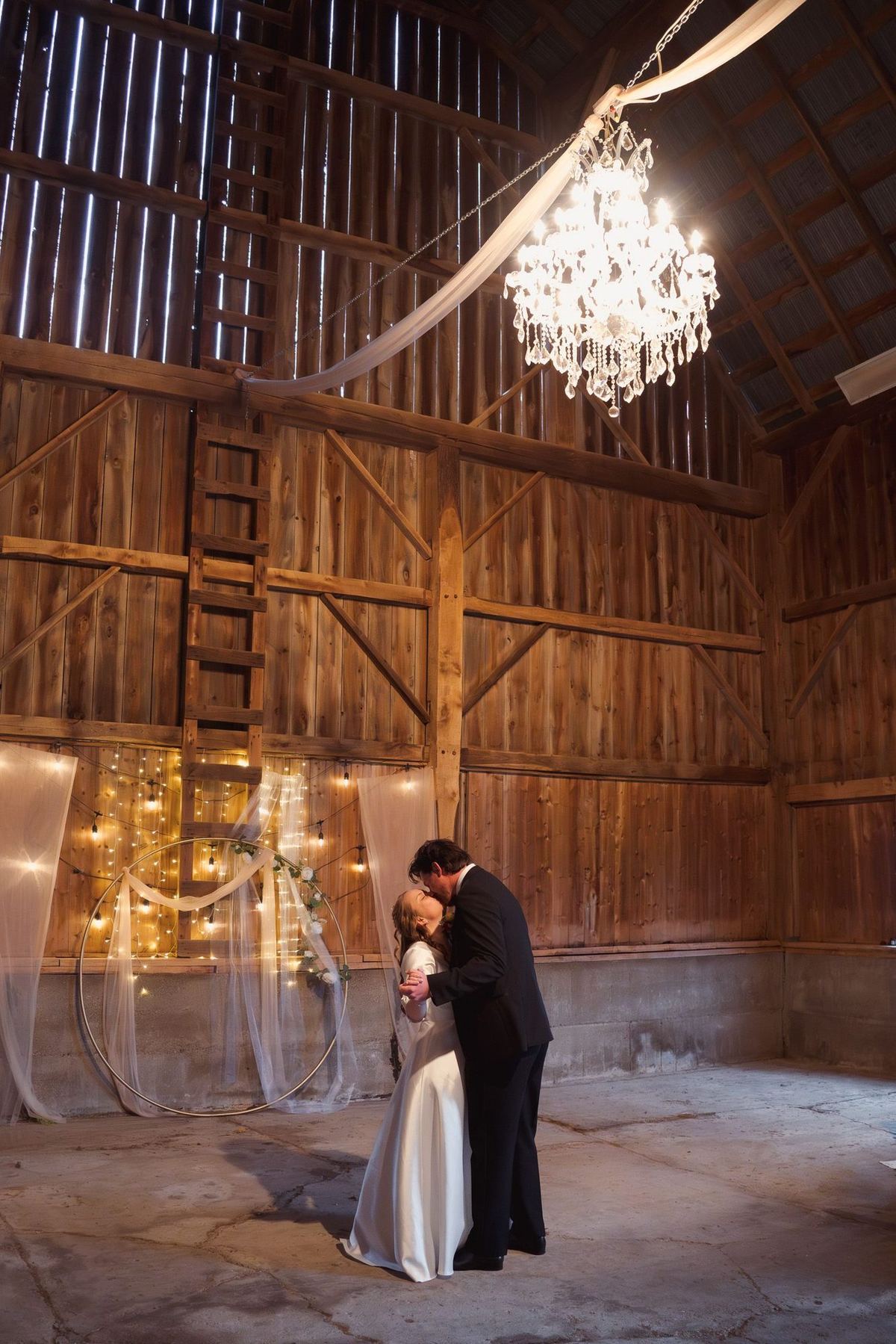 Bride and groom standing under a crystal chandelier in a rustic barn