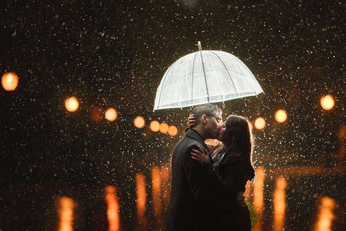 Couple kissing under a clear umbrella in the rain, backlit with string-light bokeh in a night wedding portrait