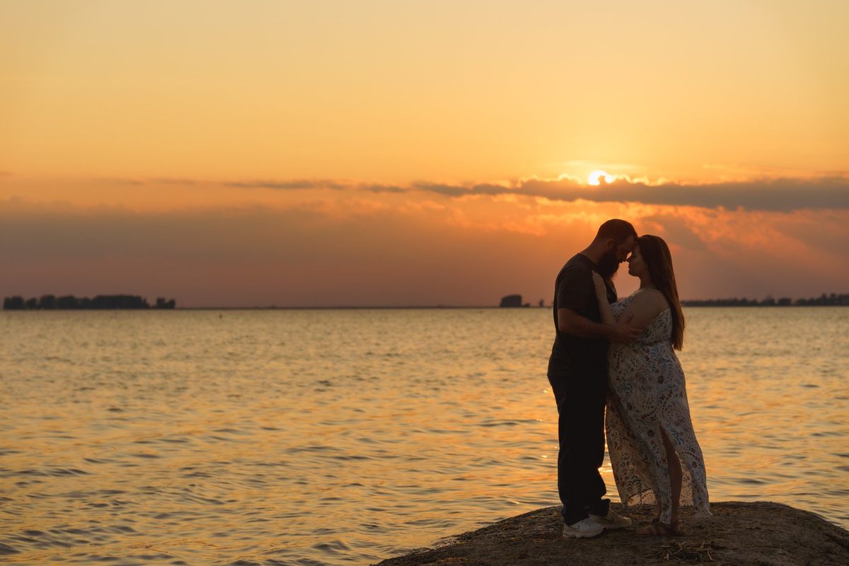 Engaged couple embracing on lakeside rocks as the sun sets over the water