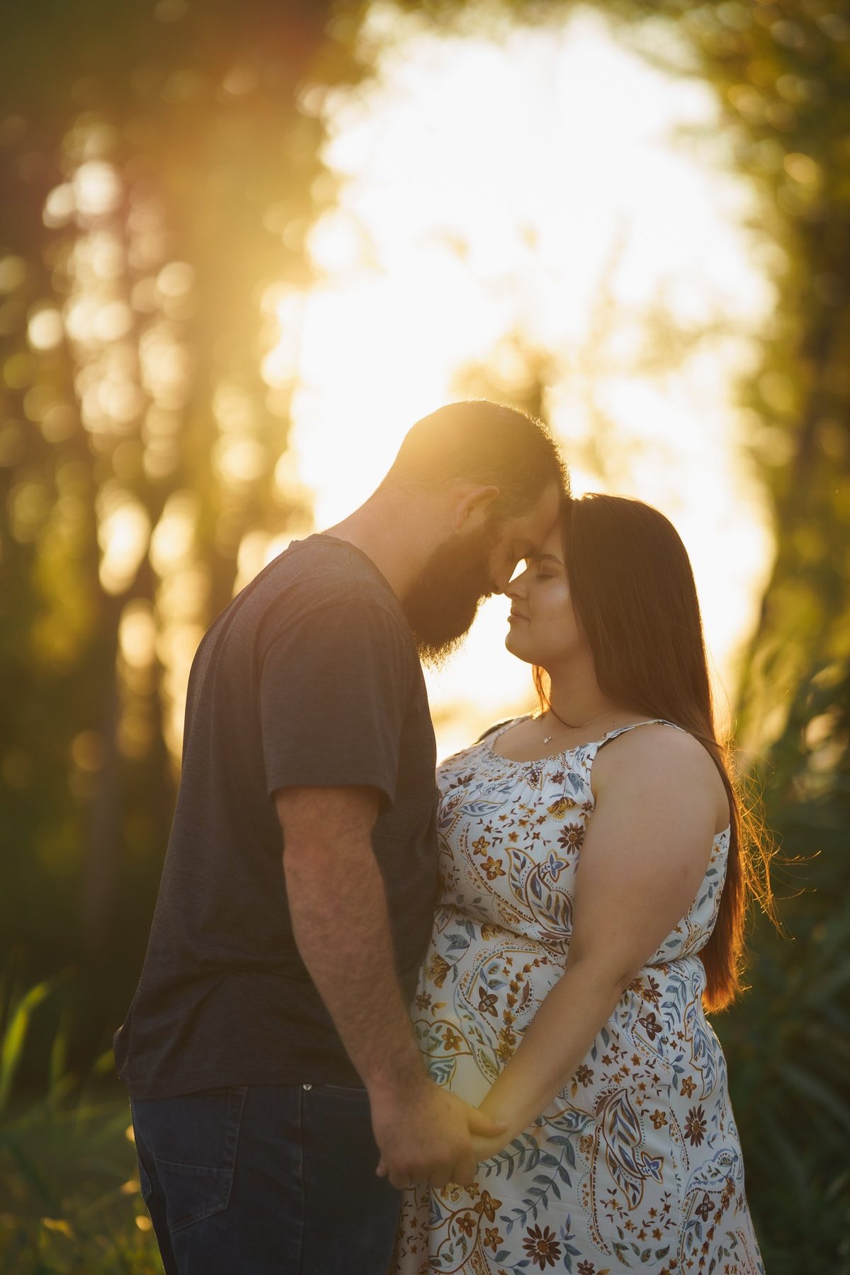Engaged couple touching foreheads among tall trees with the low sun glowing behind them