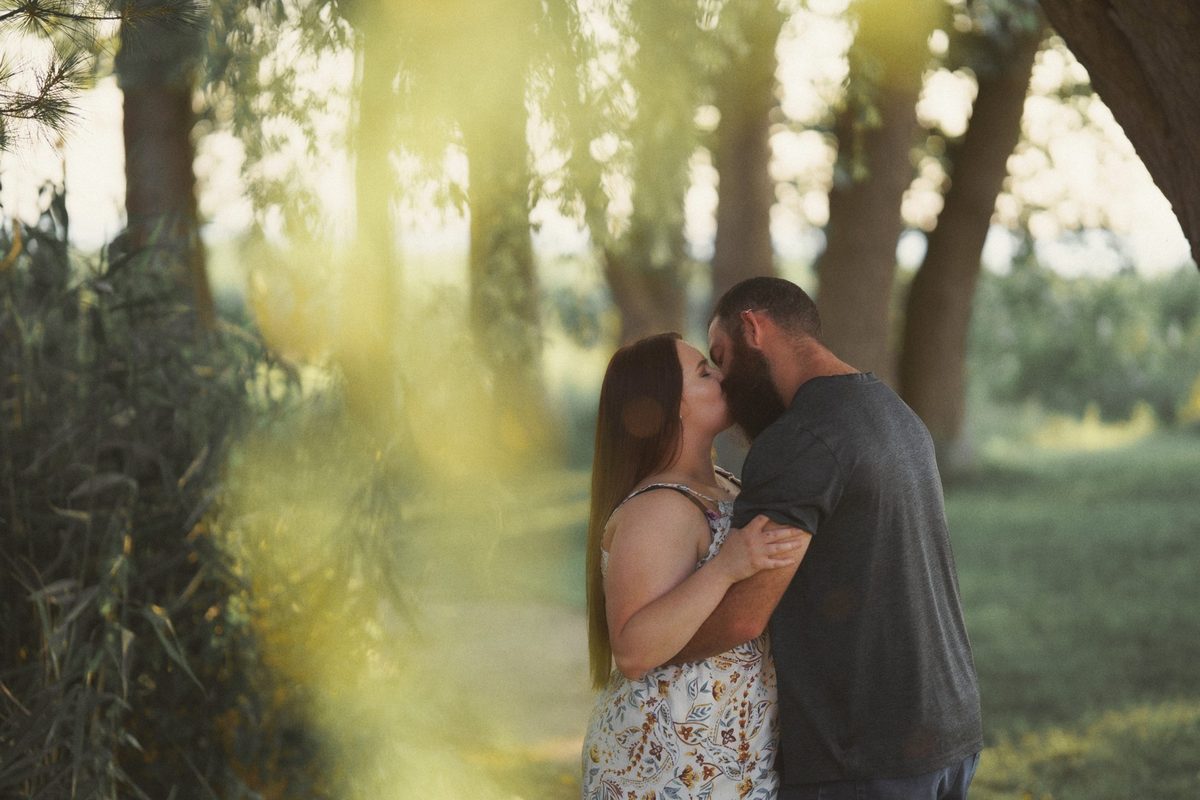 Engaged couple kissing in soft green light framed by leaves and tree trunks