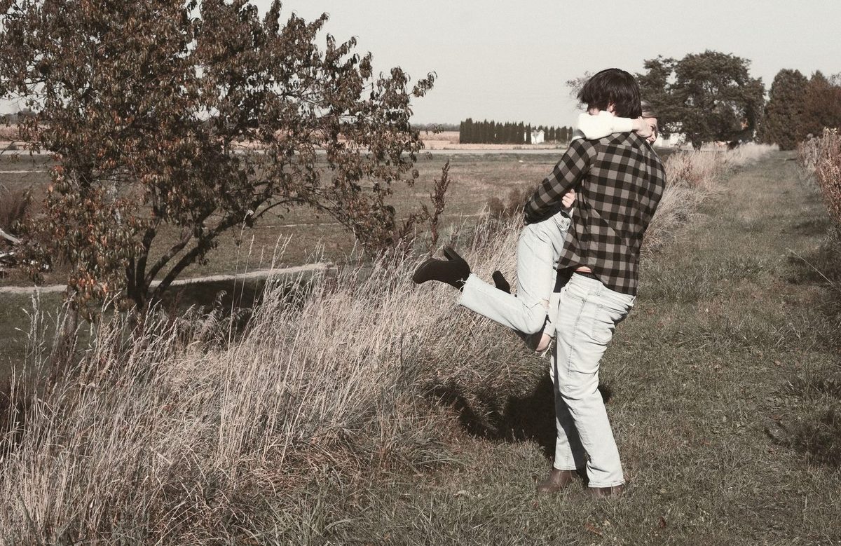 Couple goofing around in a dry grassy field, him kicking up one leg as she holds on