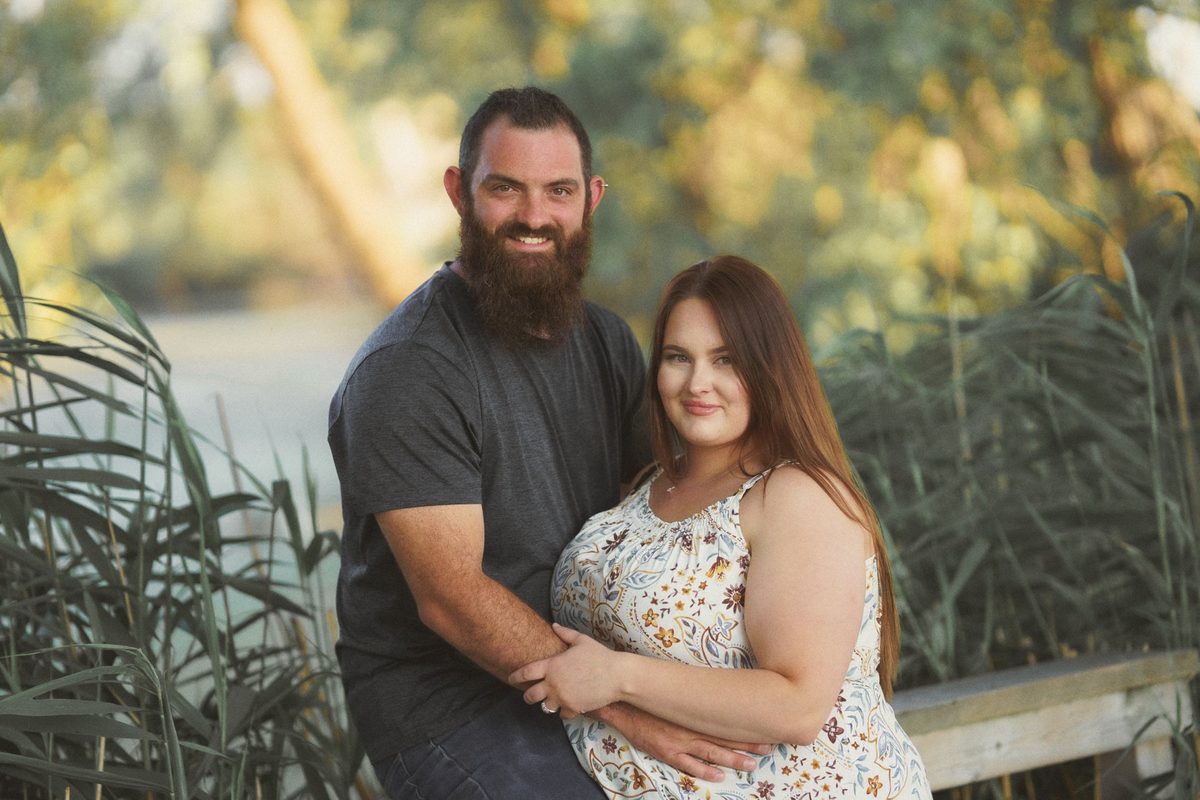 Bearded fiancé hugging his fiancée from behind beside tall reeds at Mitchell's Bay