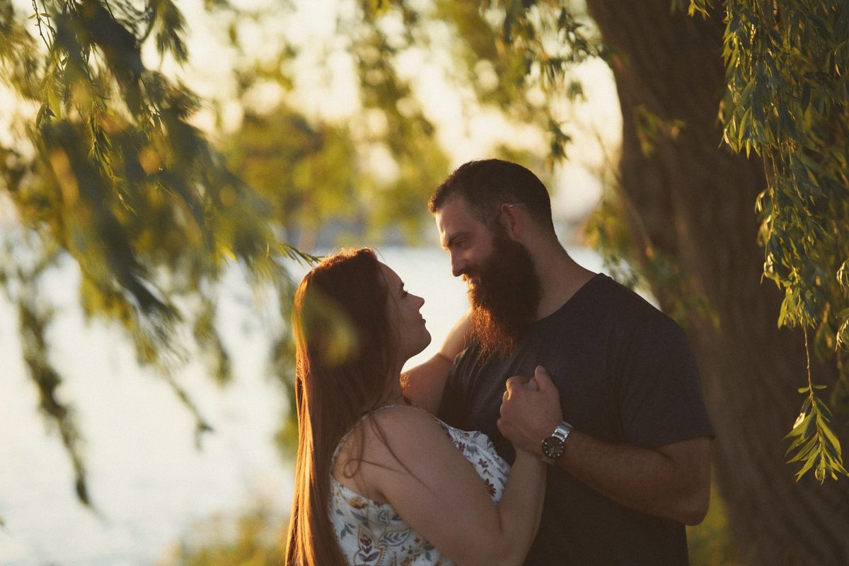 Engaged couple holding each other close beneath trailing willow branches