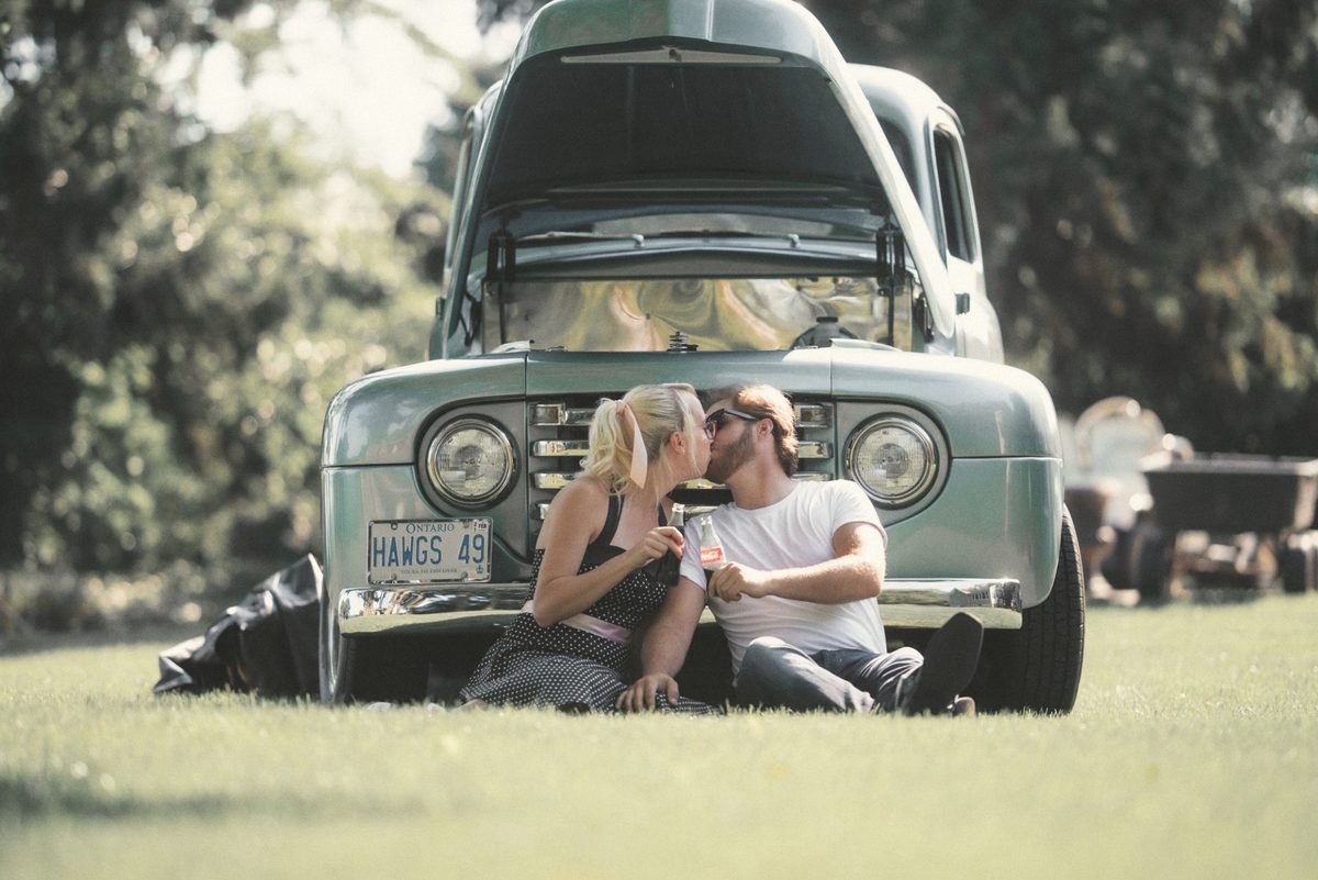 Couple sitting close on the grass in front of a vintage green pickup truck with its hood up