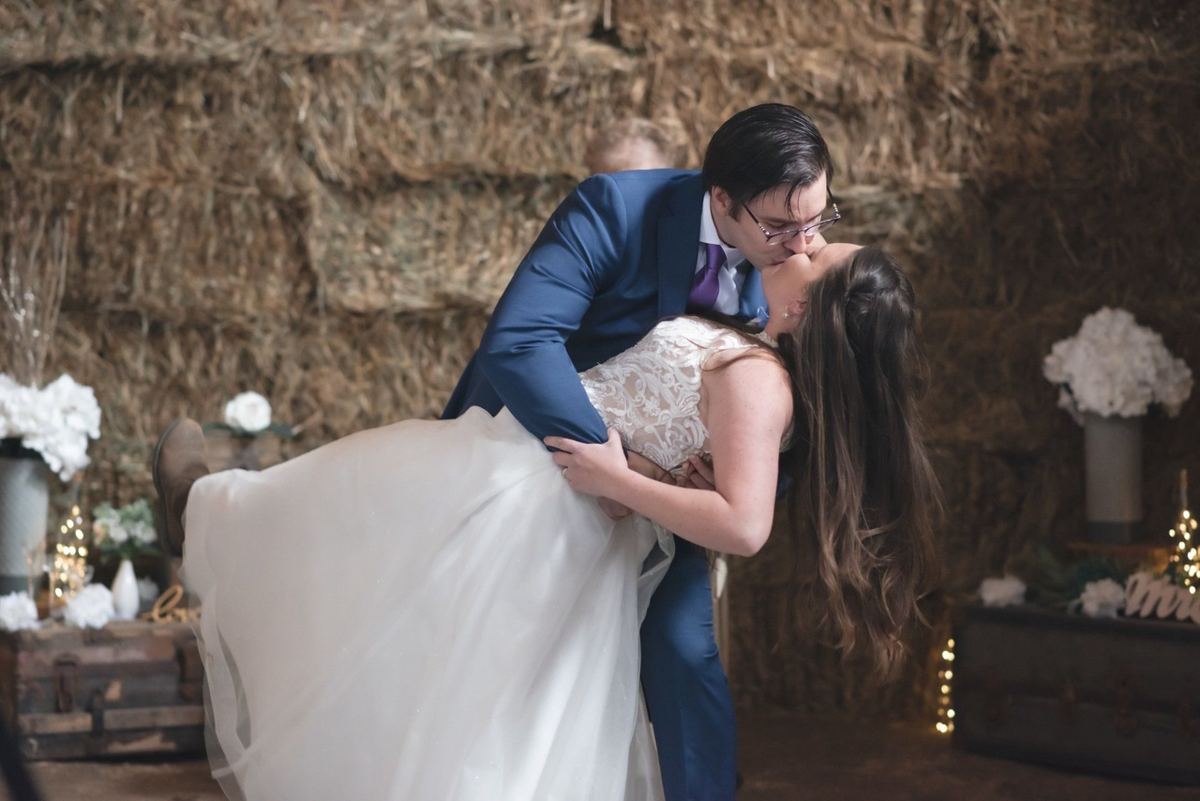 Groom dipping the bride for a kiss in a rustic barn with a chandelier and hay bales