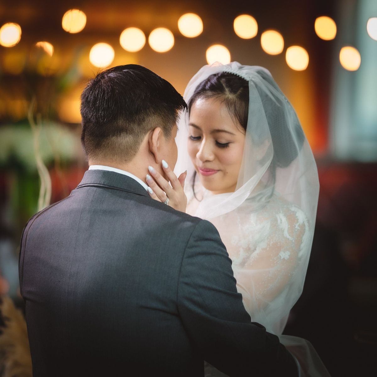 Groom holding his bride in a lace veil with warm bokeh lights behind them