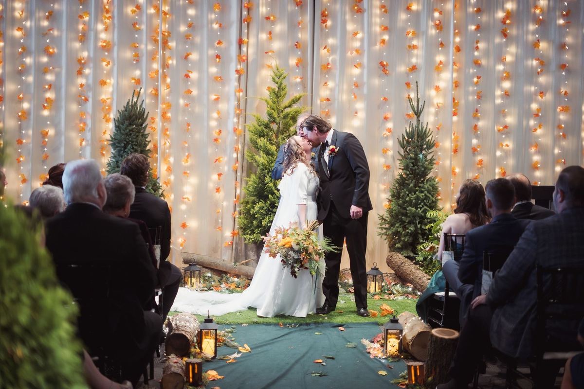 Indoor wedding ceremony under string lights with evergreen trees and guests seated