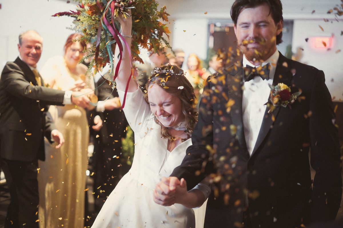 Bride laughing with her arm raised as guests throw confetti over the newlyweds after the ceremony