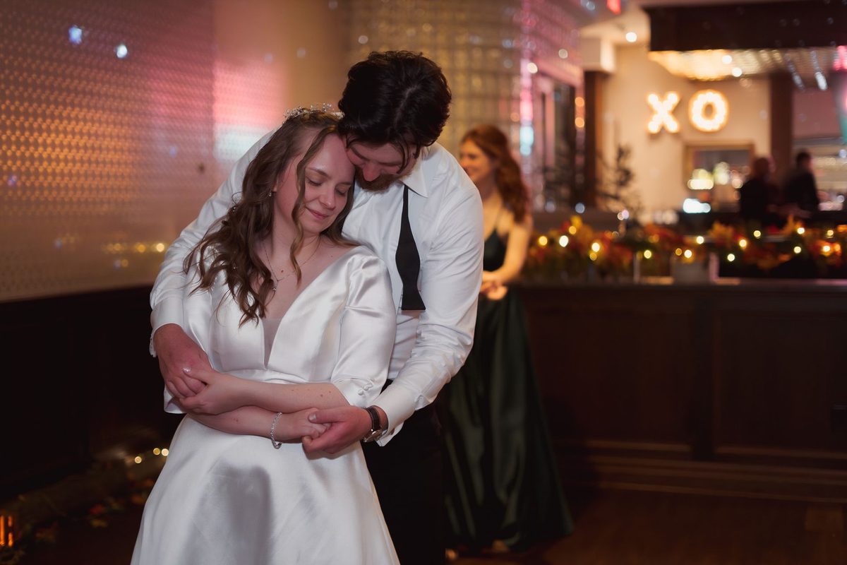 Groom hugging the bride from behind on the dance floor, her eyes closed, XO sign glowing behind them