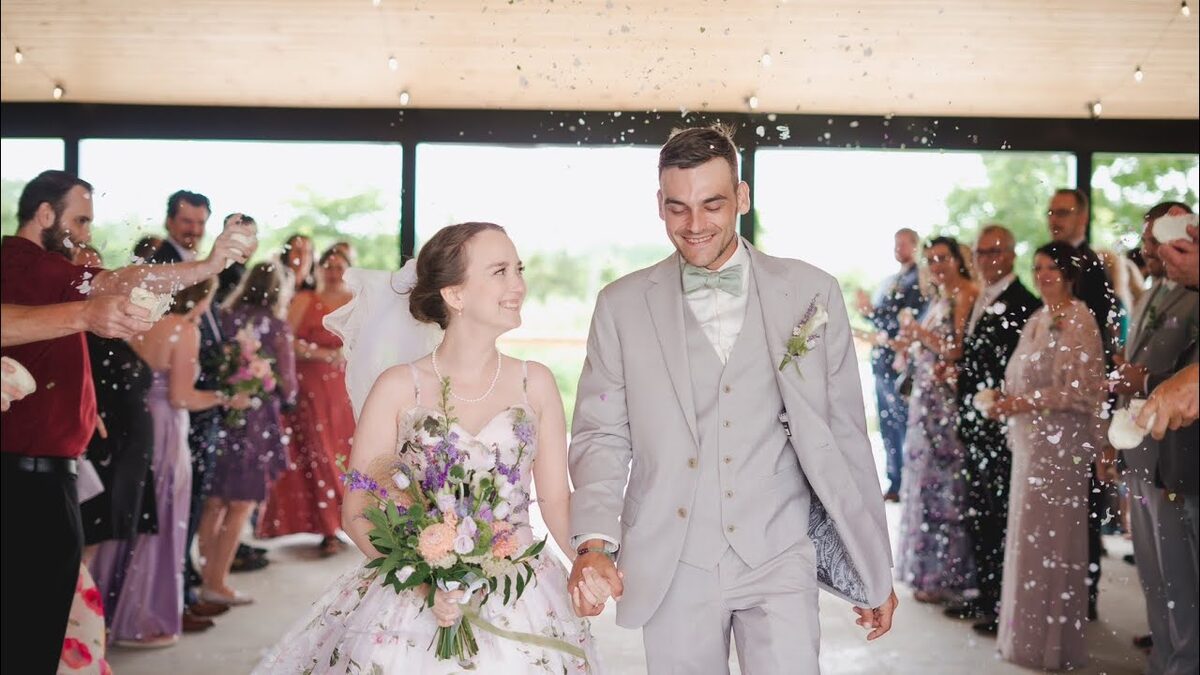 Bride and groom walking hand in hand through a confetti toss as guests cheer, a still from a wedding film