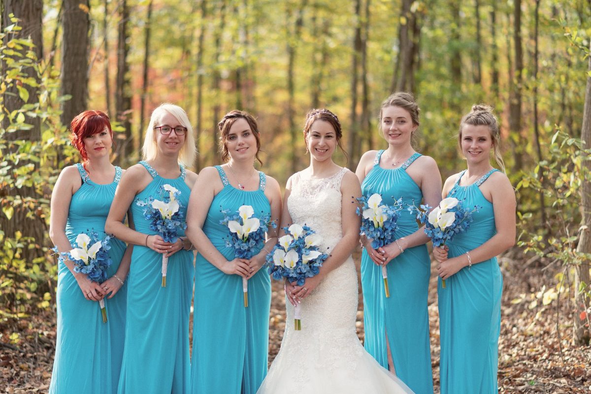 Bride with her bridesmaids in teal dresses holding blue bouquets in autumn woods