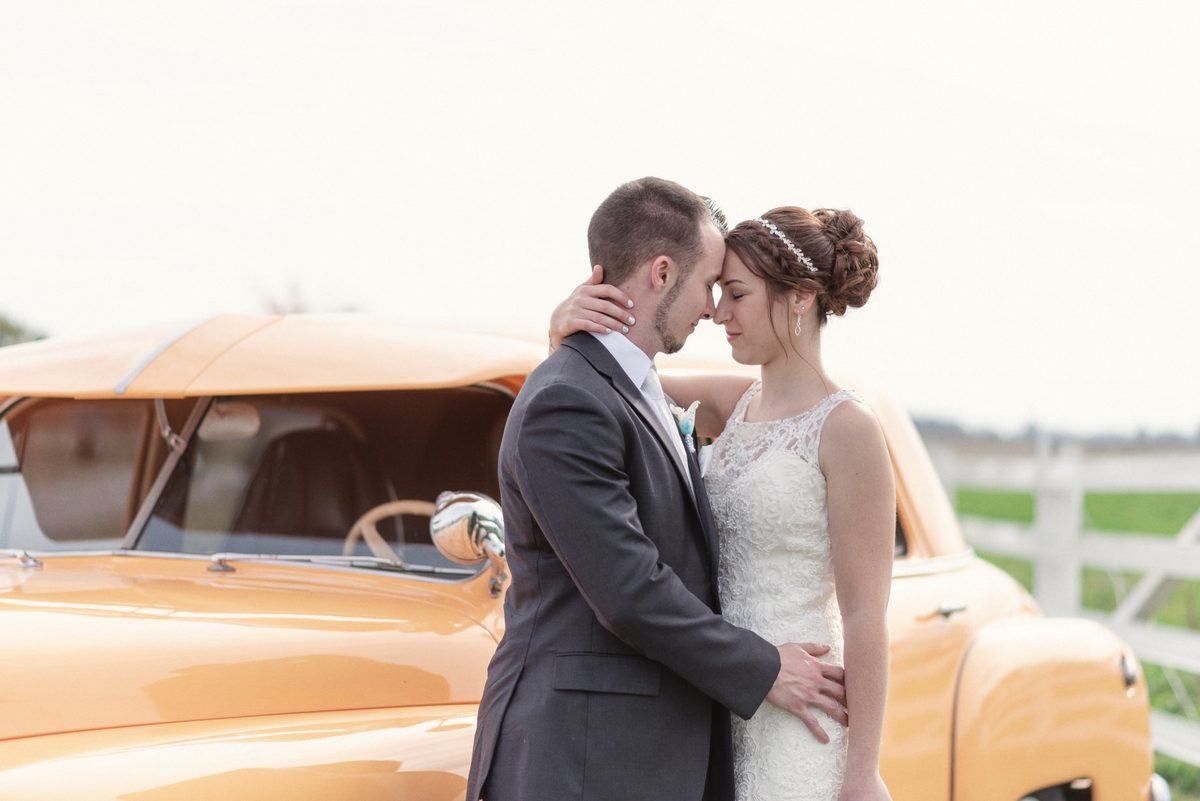 Bride and groom leaning in close beside a vintage orange car