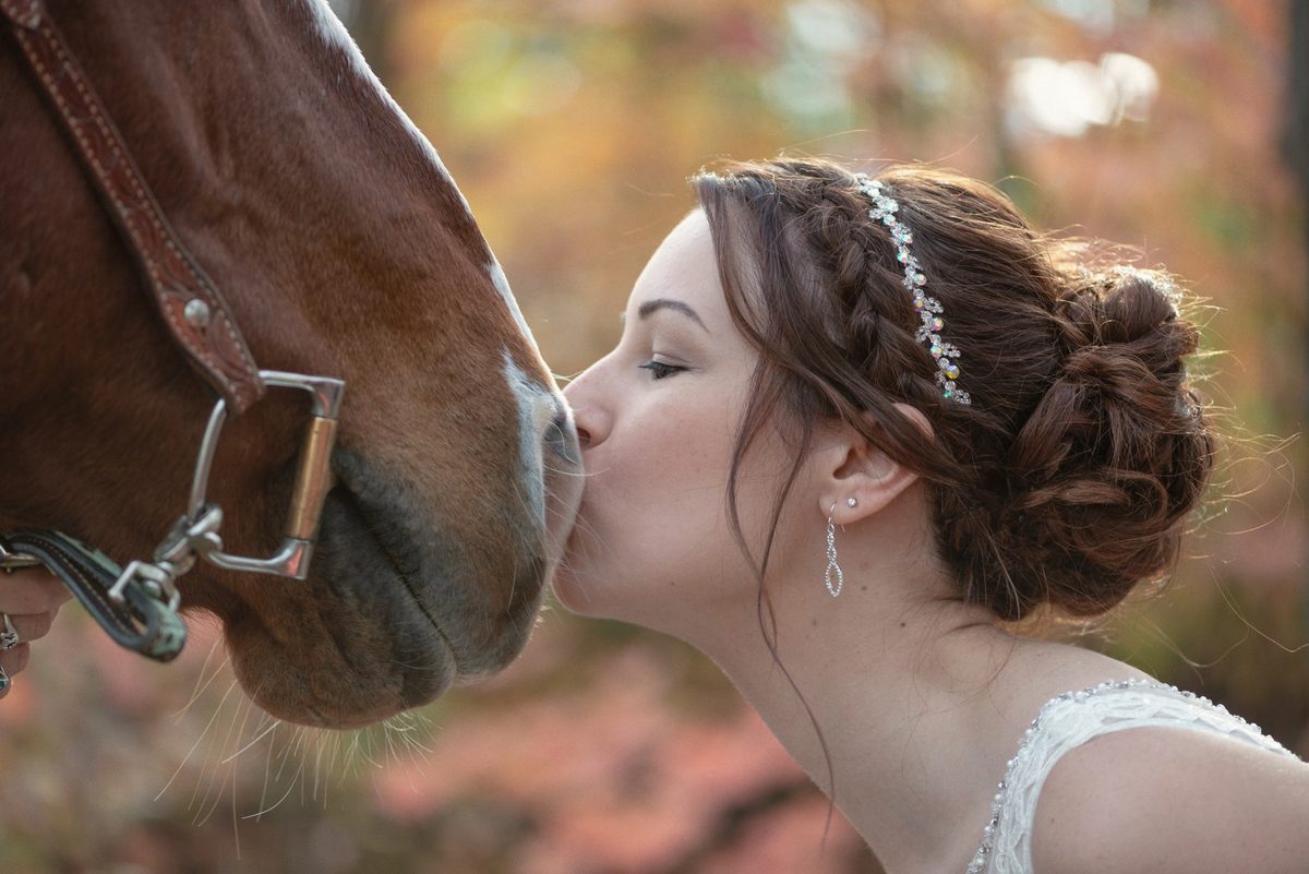 Bride leaning in to kiss a horse on the nose among autumn trees