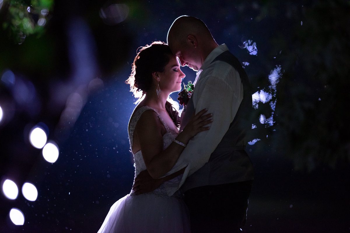Bride and groom holding each other at night, rim-lit from behind with glowing bokeh lights