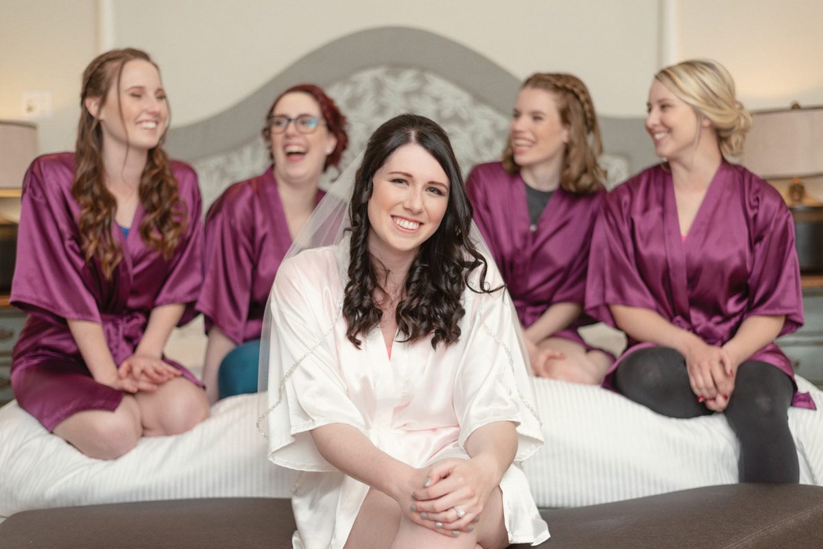 Bride in white with her bridesmaids in matching burgundy robes laughing on the bed
