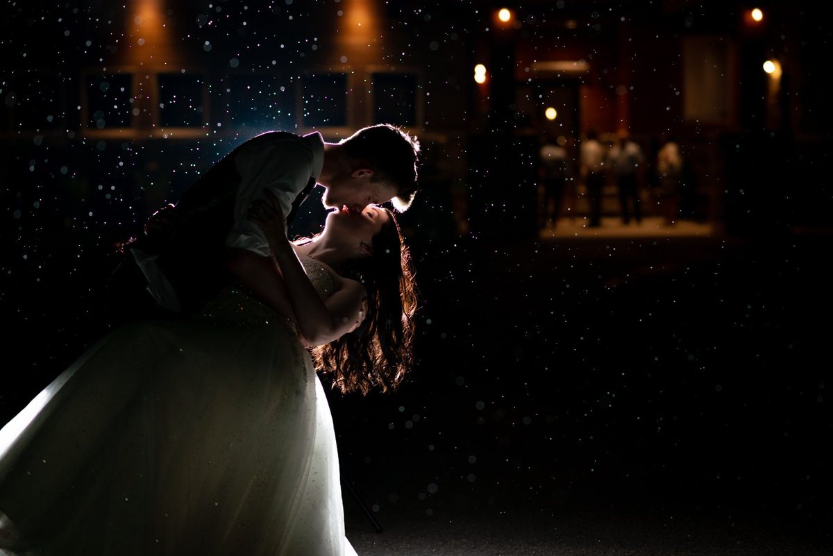 Bride and groom embracing at night, backlit by warm lights glowing behind them
