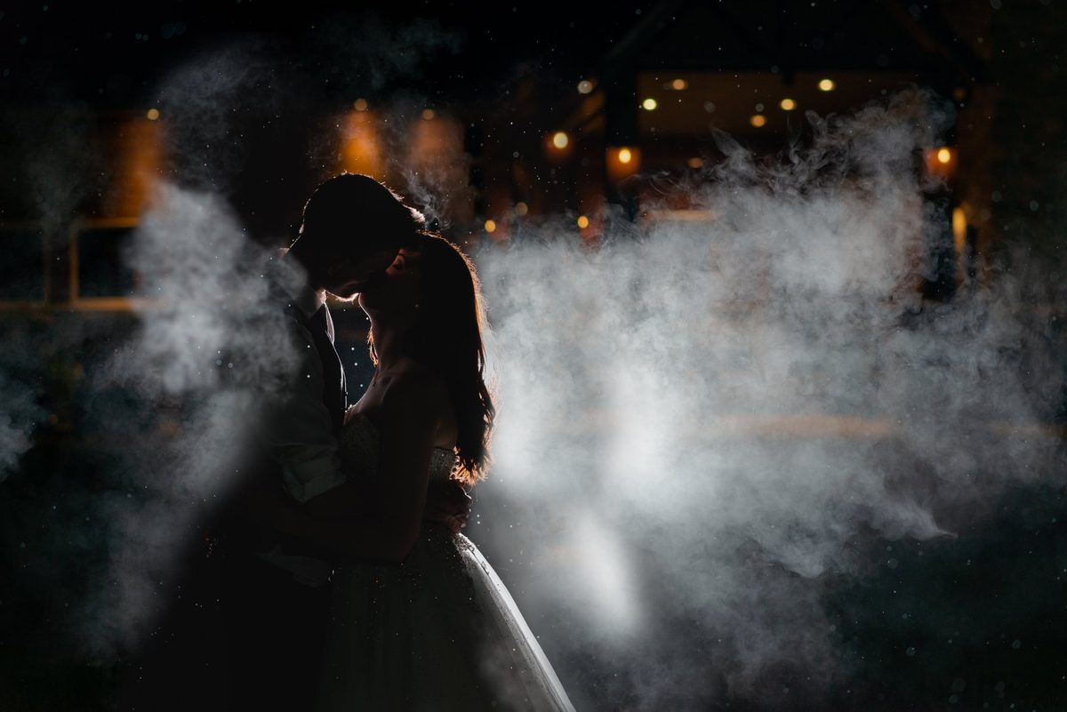 Bride and groom embracing in drifting smoke lit from behind at night