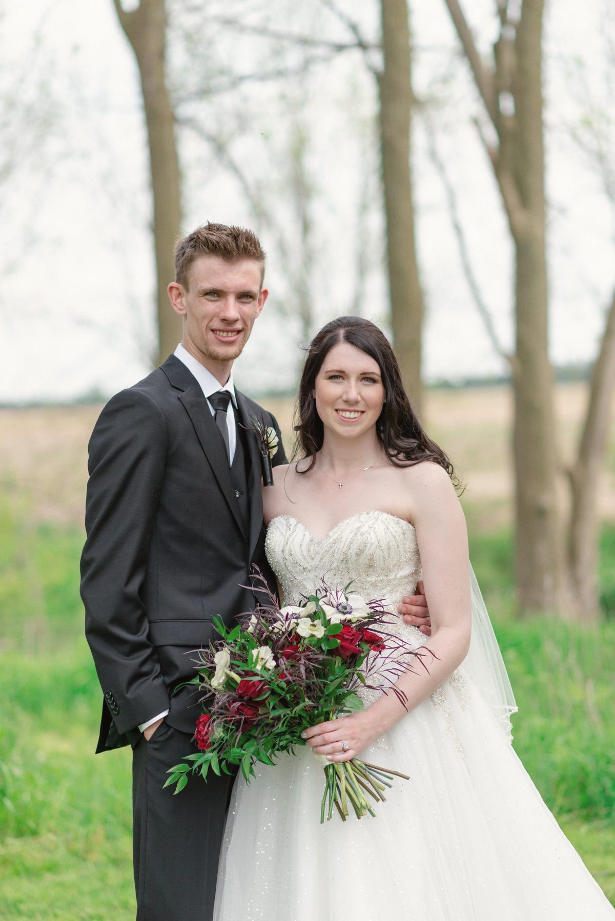 Bride and groom standing together with a bouquet of burgundy roses among spring trees