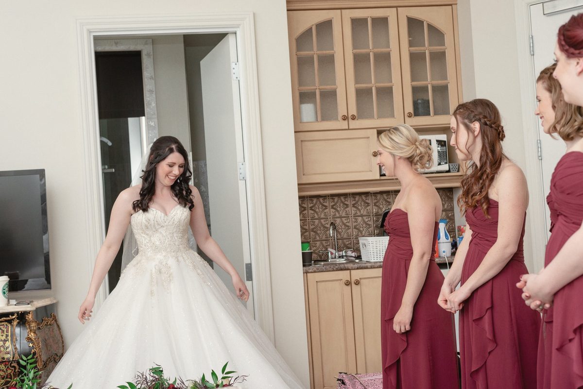 Bridesmaids in burgundy dresses watching the bride in her gown for the first time