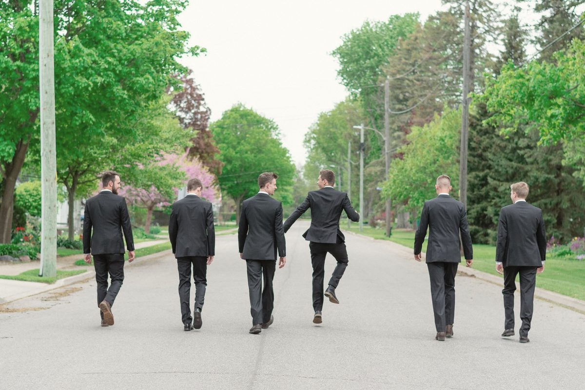 Groomsmen in dark suits walking down a tree-lined road