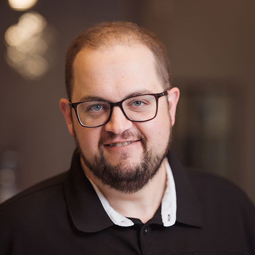 Smiling man with glasses and a beard in a dark polo, an in-office team headshot with a warm, softly blurred background for a Chatham-Kent business