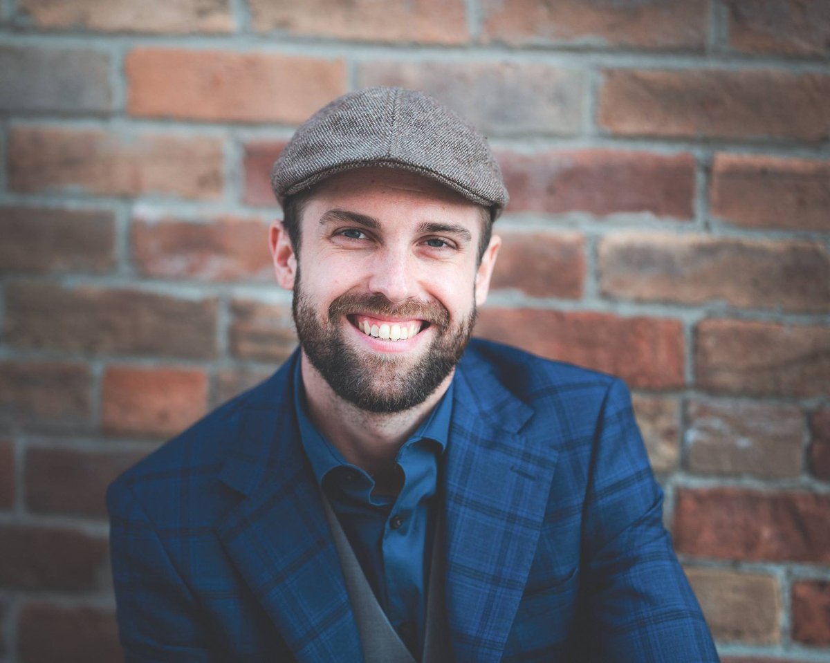 Man in a flat cap and blue blazer posing in front of a brick wall, an on-location personal brand portrait in Chatham-Kent