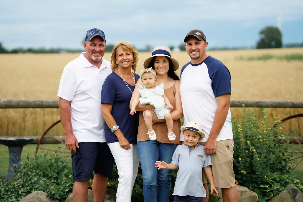 Extended family with grandparents, parents and two young kids smiling by a farm fence