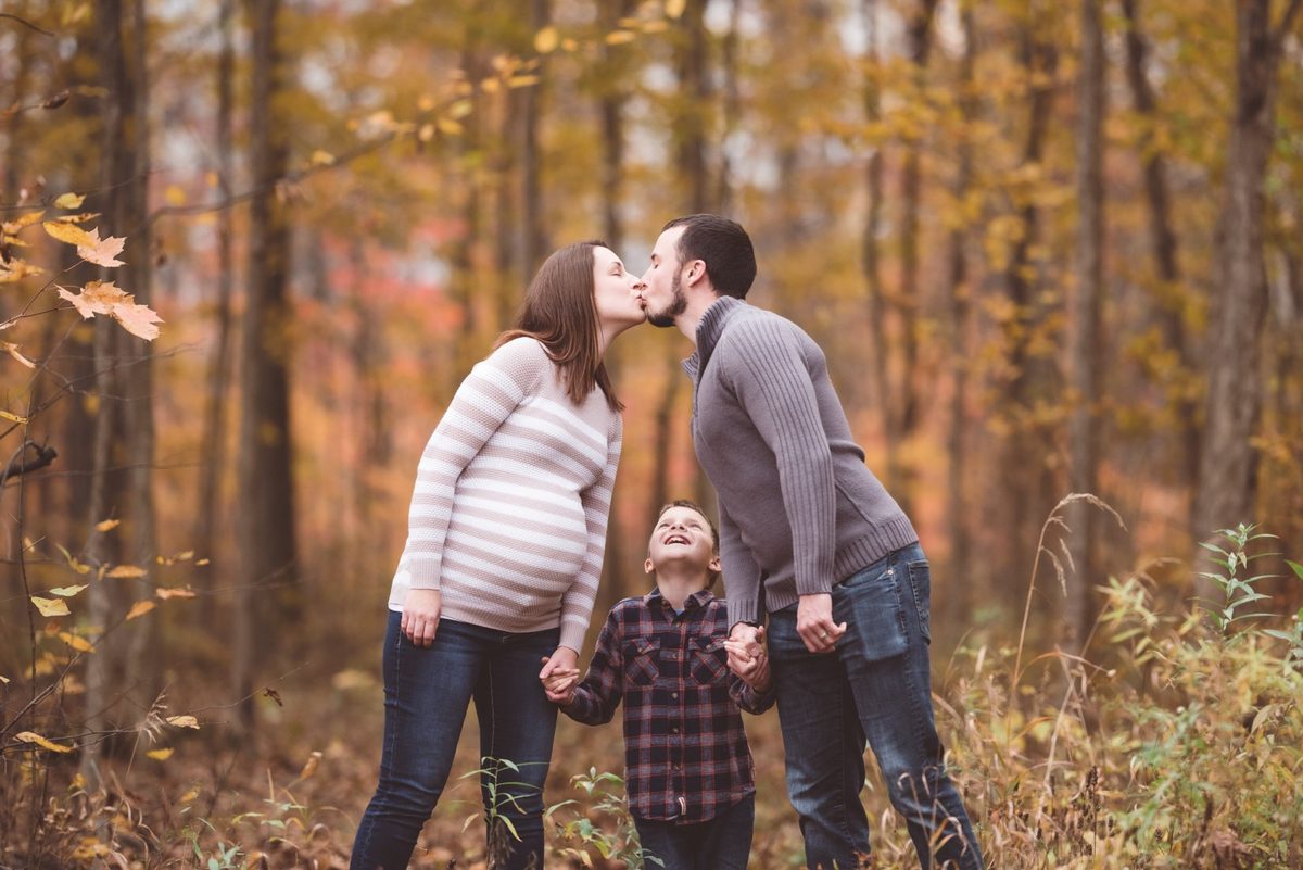 Parents sharing a kiss while holding hands with their toddler in an autumn forest