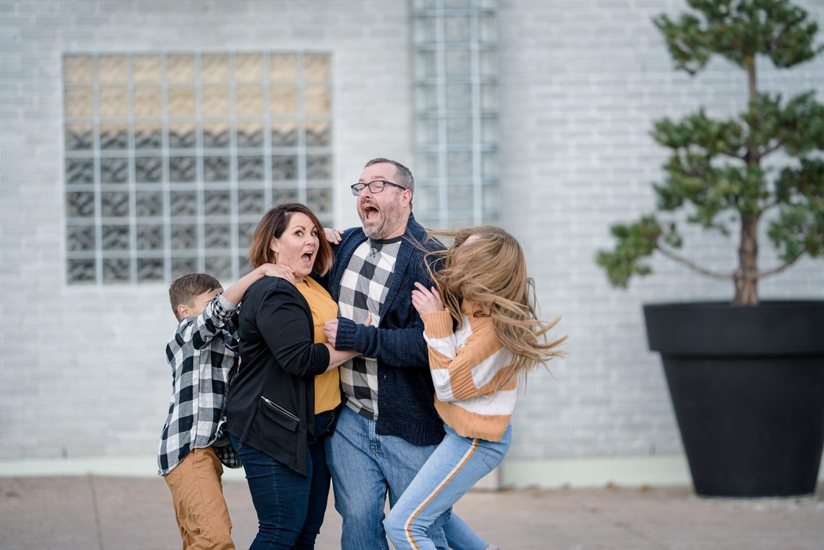 Family of five laughing and hugging in front of a white brick wall