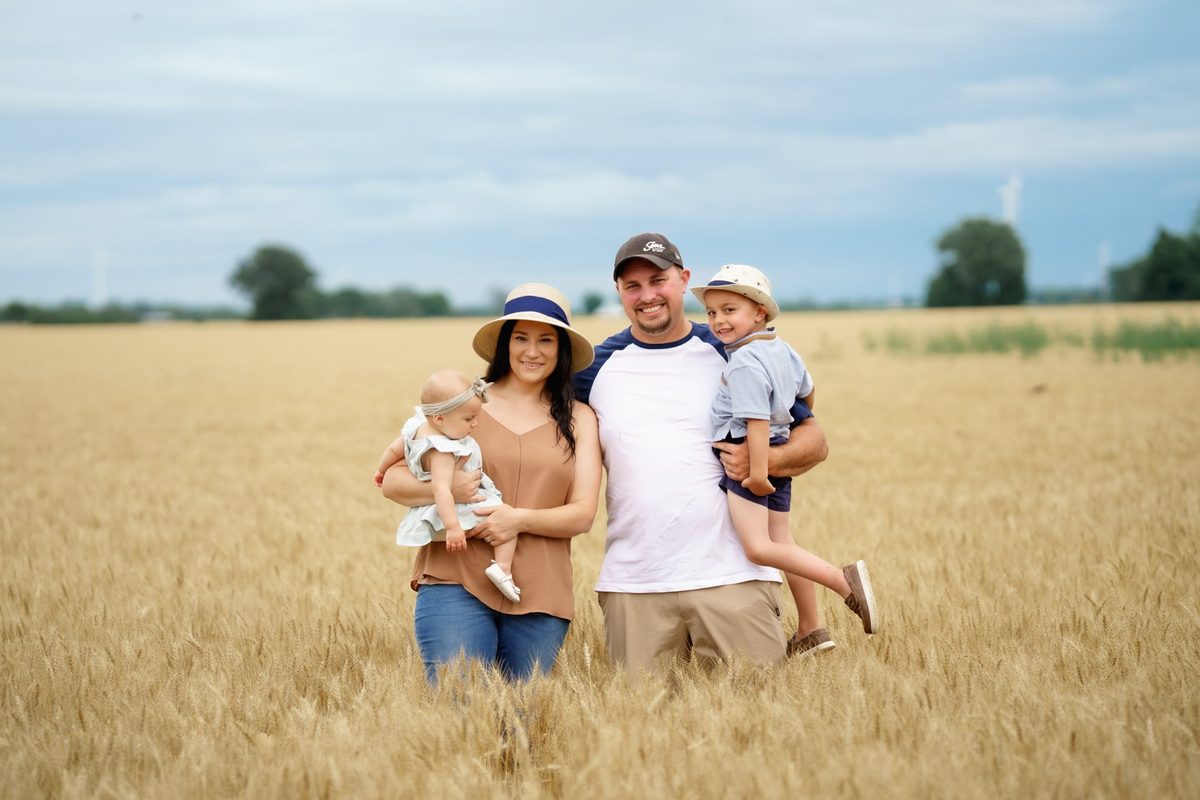 Family of five standing together in a golden wheat field, parents holding a baby and a young boy