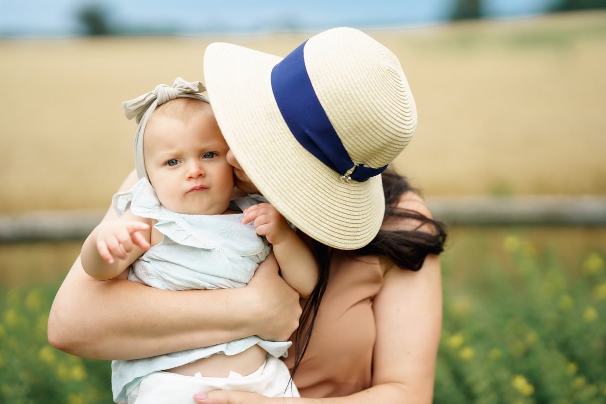 Mom in a sun hat cuddling her baby cheek to cheek in a summer field